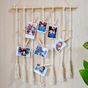 May include: A macrame photo display with wooden dowel and clips, holding several small photos. The photos are of various people. The display is hanging against a light wood-grain wall. The bottom of the display has decorative tassels.