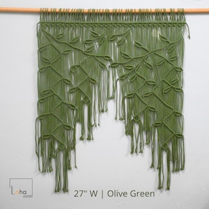 May include: An olive green macrame wall hanging suspended from a wooden rod, featuring a complex knotted vine and leaf pattern with long, loose fringe at the bottom. The text 27" W | Olive Green is printed at the bottom of the image.