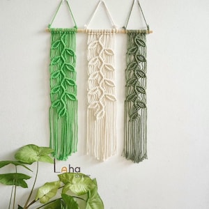 May include: Three macrame wall hangings suspended from wooden dowels, featuring leaf-shaped knot patterns in bright green, off-white, and olive green. Each piece includes a triangular cord loop for hanging and long, loose fringe at the bottom.