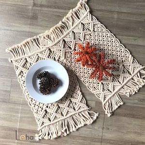 May include: A cream-colored macrame table runner featuring intricate knotted patterns and fringed edges. The runner is decorated with two orange crocheted flowers and a white plate containing three brown pinecones. The text Laha corner is visible.