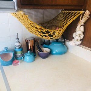 Puede incluir: Una escena de encimera de cocina con una hamaca de macram&eacute; amarilla que contiene patatas. Varios art&iacute;culos de cocina est&aacute;n colocados en la encimera, incluyendo una tetera azul, una cafetera de cobre y una botella de aceite a rayas. Un manojo de ajos cuelga a un lado.