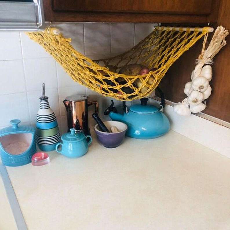 Puede incluir: Una escena de encimera de cocina con una hamaca de macram&eacute; amarilla que contiene patatas. Varios art&iacute;culos de cocina est&aacute;n colocados en la encimera, incluyendo una tetera azul, una cafetera de cobre y una botella de aceite a rayas. Un manojo de ajos cuelga a un lado.