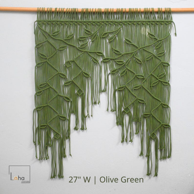 May include: An olive green macrame wall hanging suspended from a wooden rod, featuring a complex knotted vine and leaf pattern with long, loose fringe at the bottom. The text 27" W | Olive Green is printed at the bottom of the image.