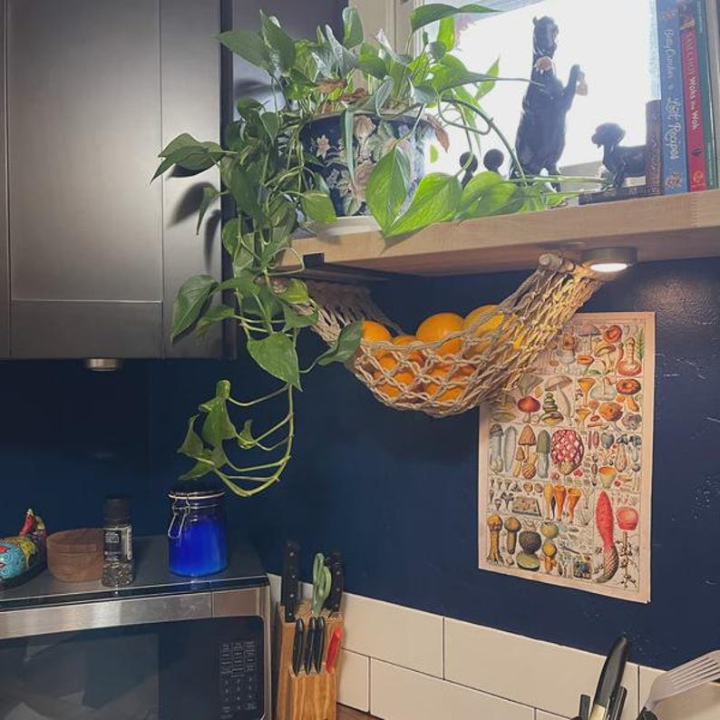Puede incluir: Interior de cocina con una cesta de frutas colgante tejida llena de naranjas. Se ven una impresi&oacute;n bot&aacute;nica y una planta en maceta. Tambi&eacute;n hay un microondas, cuchillos y otros art&iacute;culos de cocina.