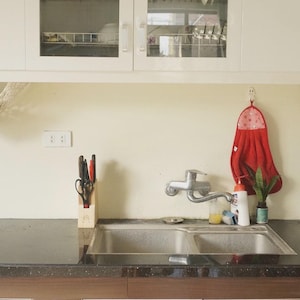 Puede incluir: Escena de cocina con un fregadero de acero inoxidable, un grifo y una toalla roja colgada en la pared. Dos hamacas de frutas de macram&eacute; cuelgan de los armarios, llenas de varias frutas. La encimera es oscura y los armarios son blancos.