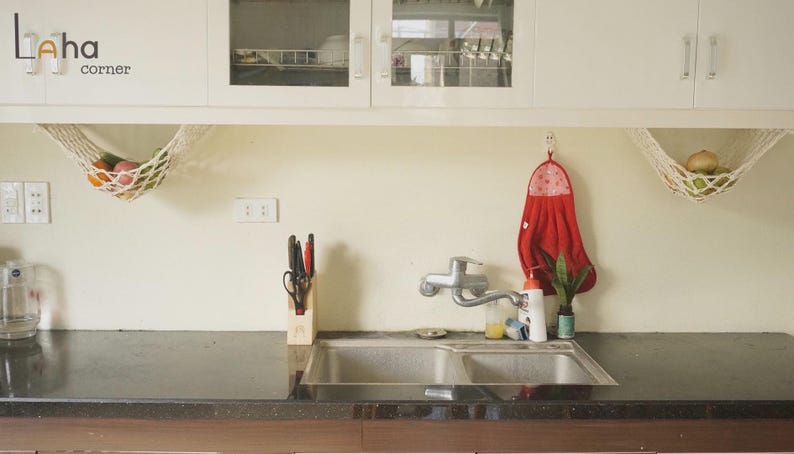 Puede incluir: Escena de cocina con un fregadero de acero inoxidable, un grifo y una toalla roja colgada en la pared. Dos hamacas de frutas de macram&eacute; cuelgan de los armarios, llenas de varias frutas. La encimera es oscura y los armarios son blancos.