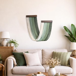 May include: A wall hanging consisting of two dark wooden bars from which long, draped cords in shades of cream, sage green, and dark forest green hang in a U-shaped pattern, creating a layered, textured fibre art display on a plain wall.