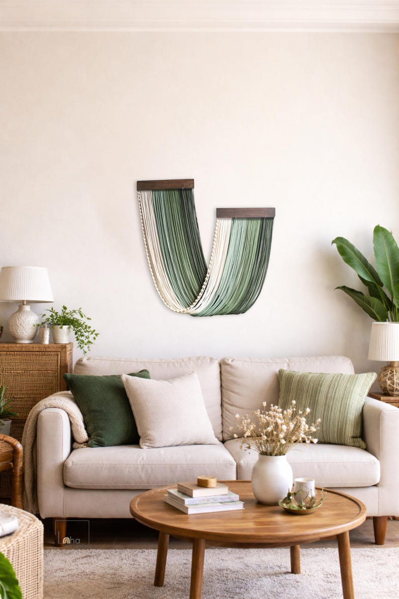 May include: A wall hanging consisting of two dark wooden bars from which long, draped cords in shades of cream, sage green, and dark forest green hang in a U-shaped pattern, creating a layered, textured fibre art display on a plain wall.
