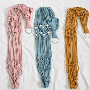 May include: Three macrame plant hangers in pink, blue, and mustard yellow. Each hanger has a wooden ring and a metal ring for hanging.