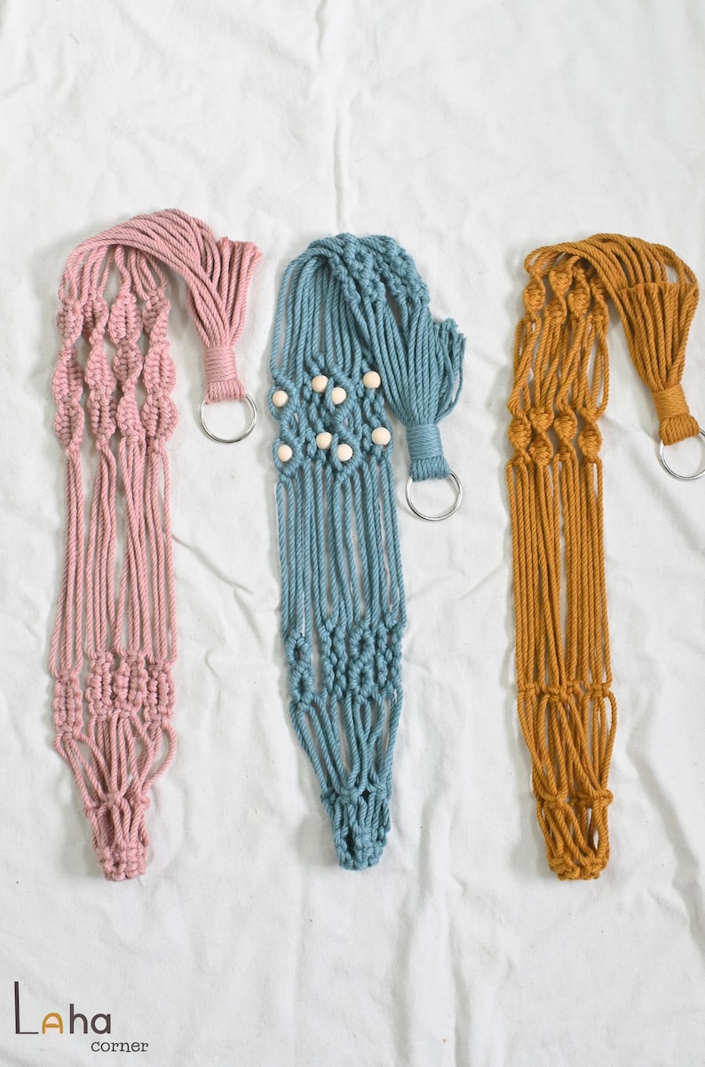May include: Three macrame plant hangers in pink, blue, and mustard yellow. Each hanger has a wooden ring and a metal ring for hanging.