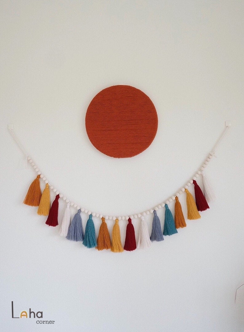 May include: A white string garland with wooden beads and colorful tassels in shades of red, yellow, blue, and white. The garland is hanging on a white wall with a large, round, orange wall hanging above it.