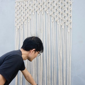 May include: A large off-white macrame wall hanging suspended from a wooden dowel. The piece features intricate knotted patterns at the top that transition into long, straight, vertical fringe strands that hang down to create a textured display.
