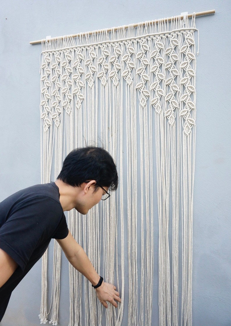 May include: A large off-white macrame wall hanging suspended from a wooden dowel. The piece features intricate knotted patterns at the top that transition into long, straight, vertical fringe strands that hang down to create a textured display.