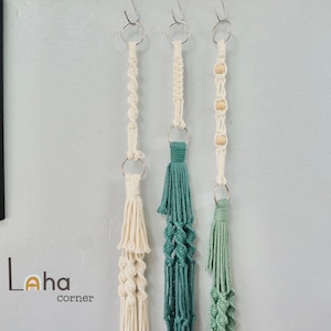 May include: Three macrame plant hangers with wooden beads hanging from hooks on a light blue wall. The hangers are made of white and green cotton cord. The text "Laha corner" is visible in the bottom left corner of the image.