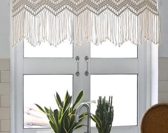 May include: An off-white macrame window valance featuring a repeating chevron pattern and long, dangling fringe with small knotted ends. The textile is suspended from a wooden rod, showcasing intricate hand-knotted cord work in a neutral tone.