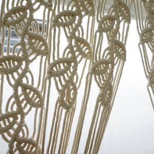 May include: An off-white macrame curtain featuring a repeating vertical pattern of knotted leaf-like shapes. The design is created with thick cotton cord, forming a series of interconnected loops and knots that hang in long, straight, vertical strands.