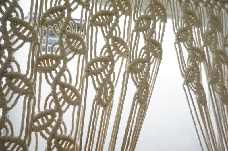 May include: An off-white macrame curtain featuring a repeating vertical pattern of knotted leaf-like shapes. The design is created with thick cotton cord, forming a series of interconnected loops and knots that hang in long, straight, vertical strands.