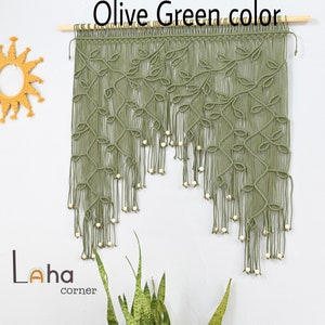 May include: An olive green macrame wall hanging suspended from a wooden dowel, featuring intricate leaf-like knot patterns and dangling light-colored wooden beads. The piece displays a cascading design with long, loose fringe ends at the bottom.
