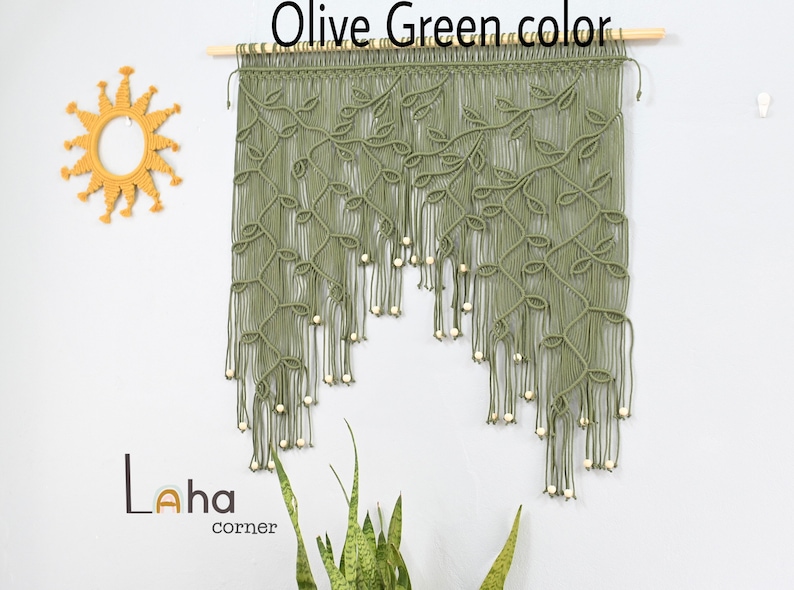 May include: An olive green macrame wall hanging suspended from a wooden dowel, featuring intricate leaf-like knot patterns and dangling light-colored wooden beads. The piece displays a cascading design with long, loose fringe ends at the bottom.
