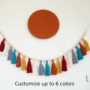 May include: A colorful tassel garland with wooden beads, featuring red, white, blue, teal, orange, and yellow tassels. The garland is hanging on a white wall with a large orange circle behind it. The text "Customize up to 6 colors" is below the garland. The text "Laha corner" is in the bottom right corner of the image.