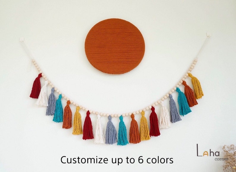 May include: A colorful tassel garland with wooden beads, featuring red, white, blue, teal, orange, and yellow tassels. The garland is hanging on a white wall with a large orange circle behind it. The text "Customize up to 6 colors" is below the garland. The text "Laha corner" is in the bottom right corner of the image.