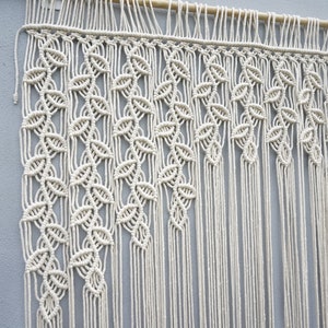 May include: A cream-colored macrame wall hanging suspended from a wooden dowel. The textile features intricate knotted patterns forming vertical leaf-like shapes that transition into long, straight, unknotted fringe strands at the bottom.