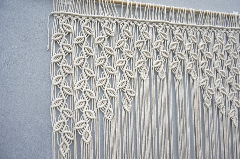 May include: A cream-colored macrame wall hanging suspended from a wooden dowel. The textile features intricate knotted patterns forming vertical leaf-like shapes that transition into long, straight, unknotted fringe strands at the bottom.