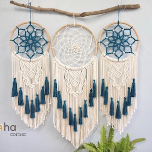 May include: Three macrame wall hangings suspended from a horizontal wooden branch. Two outer pieces feature dark teal geometric patterns in wooden hoops, while the center piece has a cream web design. All include cream fringe and teal tassels.