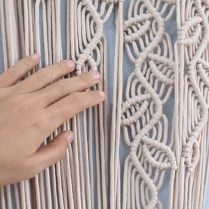 May include: A beige macrame wall hanging featuring intricate knotted patterns and long, vertical hanging cords. The design includes repeating leaf-like shapes created with tight knots. The logo Lana corner is visible in the bottom left corner.