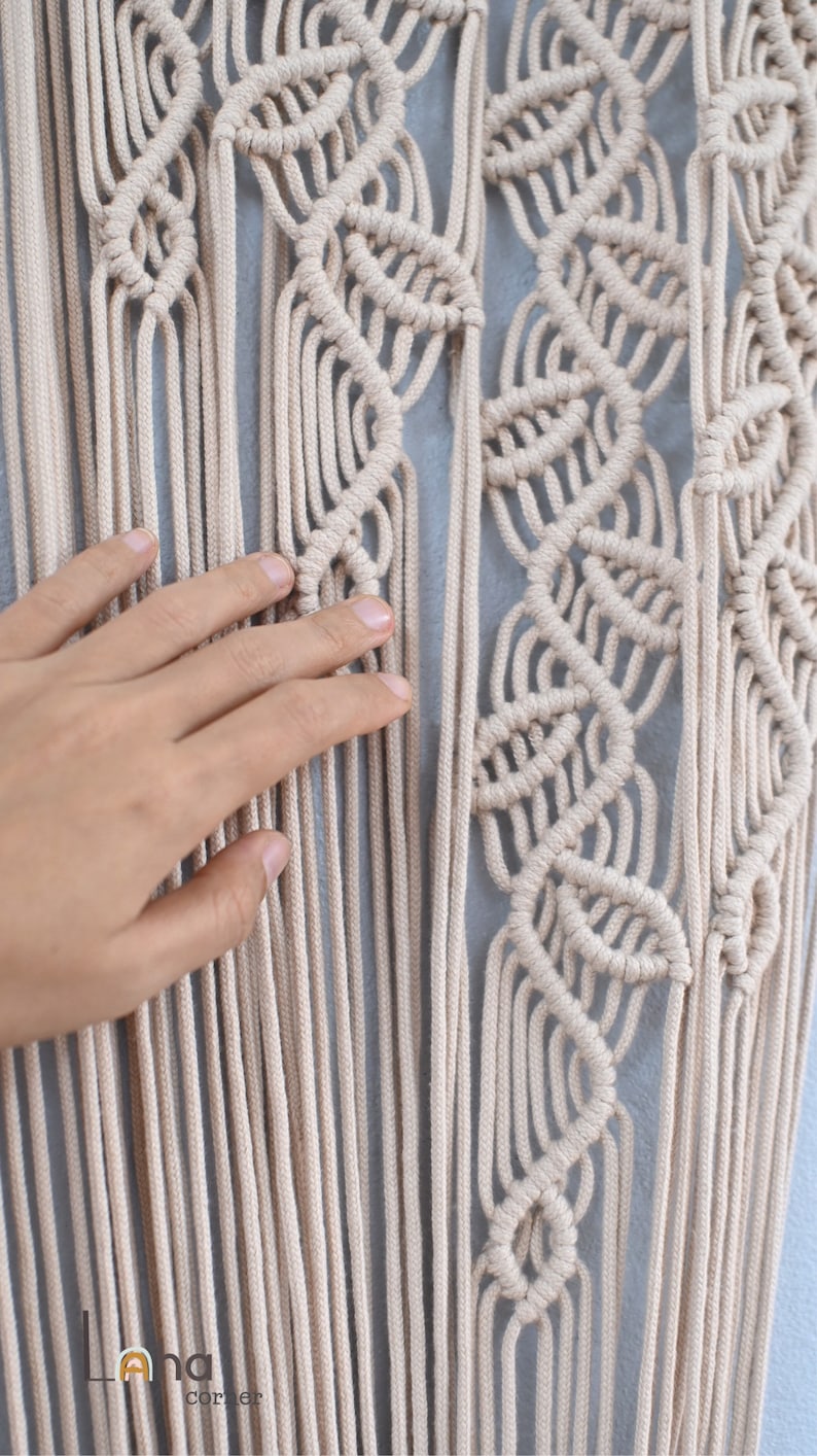 May include: A beige macrame wall hanging featuring intricate knotted patterns and long, vertical hanging cords. The design includes repeating leaf-like shapes created with tight knots. The logo Lana corner is visible in the bottom left corner.