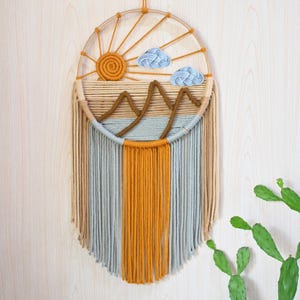 Könnte beinhalten: Handgefertigter Makramee-Wandbehang mit Sonne, Wolken und Bergen. Das Design verwendet Gelb-, Braun- und Blautöne. Der untere Teil hat lange, vertikale Fransen in verschiedenen Farben. Ein dekoratives Element für die Inneneinrichtung.
