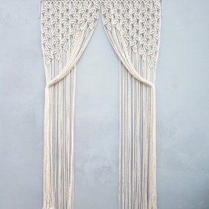 May include: An off-white macrame curtain hanging from a wooden rod. The upper section features a dense, repeating leaf-like knot pattern, while the lower section consists of long, vertical, unknotted fringe strands that are gathered at the sides.