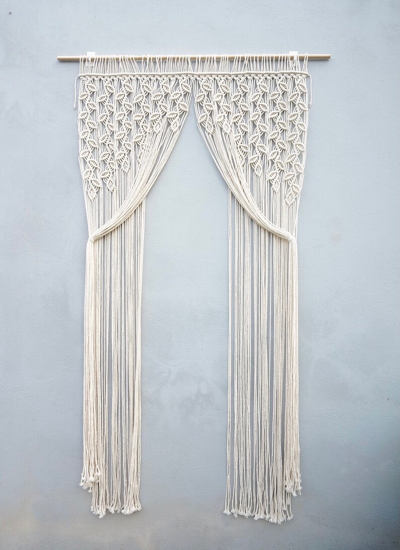 May include: An off-white macrame curtain hanging from a wooden rod. The upper section features a dense, repeating leaf-like knot pattern, while the lower section consists of long, vertical, unknotted fringe strands that are gathered at the sides.