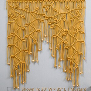 May include: A mustard yellow macrame wall hanging featuring a leaf-like pattern created with intricate knotted cords, suspended from a wooden dowel. The piece includes light wooden beads at the ends of the fringe and text stating: Shown in: 30" W x 35" L | Mustard. Other sizes and colors are available.