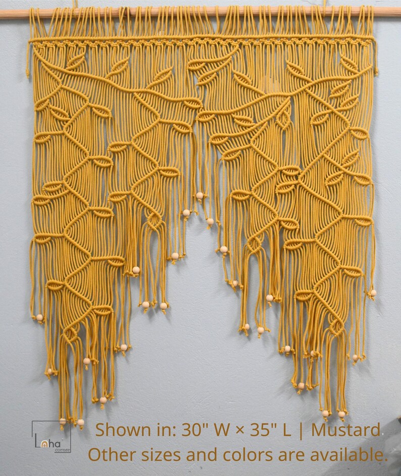 May include: A mustard yellow macrame wall hanging featuring a leaf-like pattern created with intricate knotted cords, suspended from a wooden dowel. The piece includes light wooden beads at the ends of the fringe and text stating: Shown in: 30" W x 35" L | Mustard. Other sizes and colors are available.