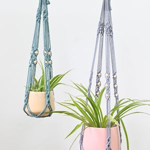 May include: Two macrame plant hangers, one in light blue and one in gray, with wooden beads. Each hanger is holding a potted plant.