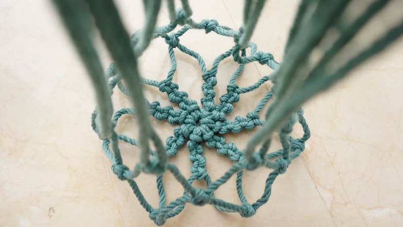 Pu&ograve; includere: Un portavasi sospeso in macram&egrave; blu-verde con un motivo a stella. Il macram&egrave; &egrave; realizzato con una corda spessa e presenta un design annodato.