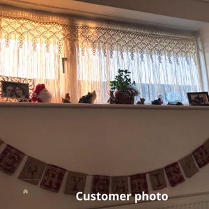 May include: A cream-colored macrame valance with a geometric knotted pattern hangs over a window. Below, a banner made of burlap and red plaid fabric squares displays the text MERRY CHRISTMAS, with each letter printed on a separate fabric panel.