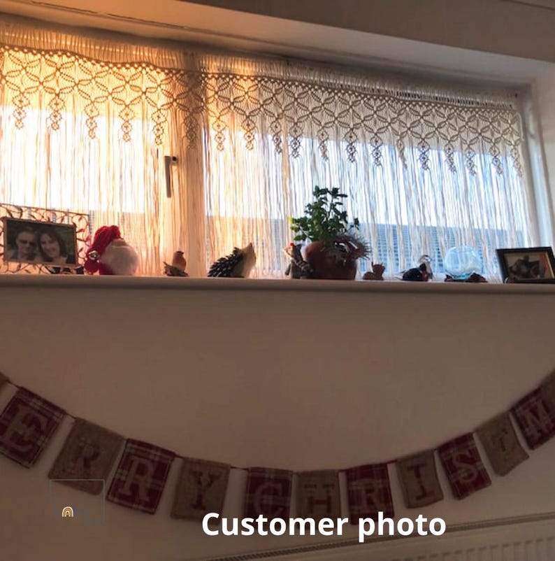 May include: A cream-colored macrame valance with a geometric knotted pattern hangs over a window. Below, a banner made of burlap and red plaid fabric squares displays the text MERRY CHRISTMAS, with each letter printed on a separate fabric panel.