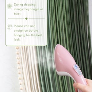 May include: A handheld pink garment steamer emitting steam onto a wall hanging made of long, vertical green and cream-coloured strings attached to a wooden bar. A white informational box with green text advises to iron and straighten strings.