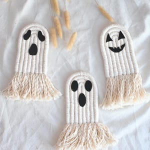May include: Three handmade macrame ghost decorations with black facial features and fringed bottoms. The ghosts are white and beige, arranged on a white fabric background, perfect for Halloween.