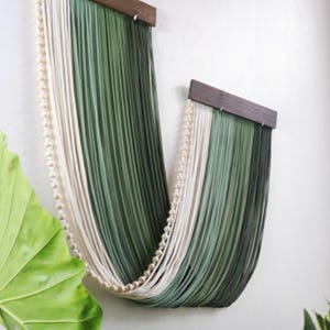 May include: A wall hanging featuring two dark brown wooden bars from which long, draped strands of cream and forest green yarn are suspended. A single string of small, round, light-coloured wooden beads follows the curve of the yarn strands.