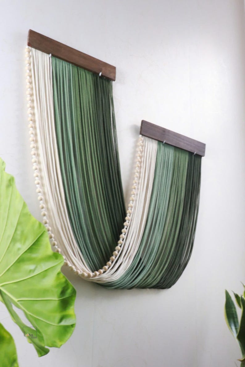 May include: A wall hanging featuring two dark brown wooden bars from which long, draped strands of cream and forest green yarn are suspended. A single string of small, round, light-coloured wooden beads follows the curve of the yarn strands.