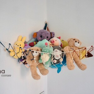 May include: A white string hammock with wooden beads hangs from the corner of a white wall. The hammock is filled with stuffed animals, including a purple elephant, a green dinosaur, a pink pig, a brown bear, and a yellow bunny. The text "Laha corner" is printed on the wall below the hammock.