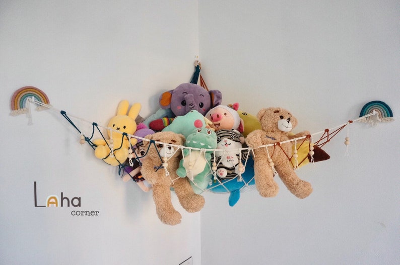 May include: A white string hammock with wooden beads hangs from the corner of a white wall. The hammock is filled with stuffed animals, including a purple elephant, a green dinosaur, a pink pig, a brown bear, and a yellow bunny. The text "Laha corner" is printed on the wall below the hammock.
