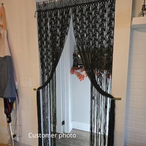 May include: A black macrame doorway curtain featuring an intricate knotted leaf pattern across the top section, transitioning into long, straight hanging fringe strands that are gathered and held back on each side of the door frame.