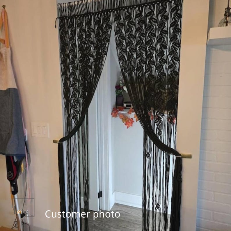 May include: A black macrame doorway curtain featuring an intricate knotted leaf pattern across the top section, transitioning into long, straight hanging fringe strands that are gathered and held back on each side of the door frame.