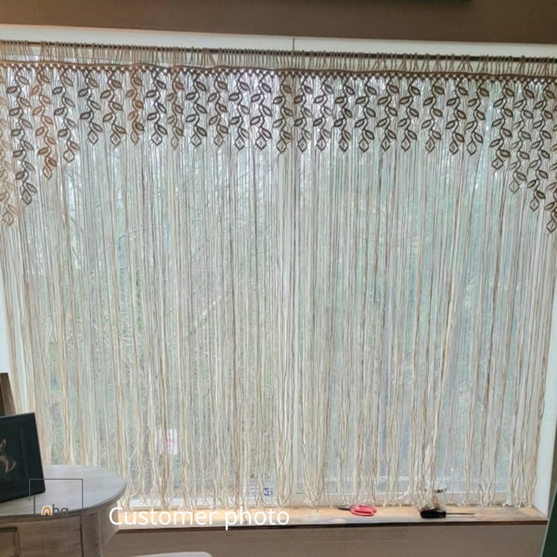 May include: A cream-colored macrame window curtain featuring a decorative top border with a repeating leaf pattern. The curtain consists of numerous long, vertical hanging cords that extend downward, creating a fringe effect across the window.
