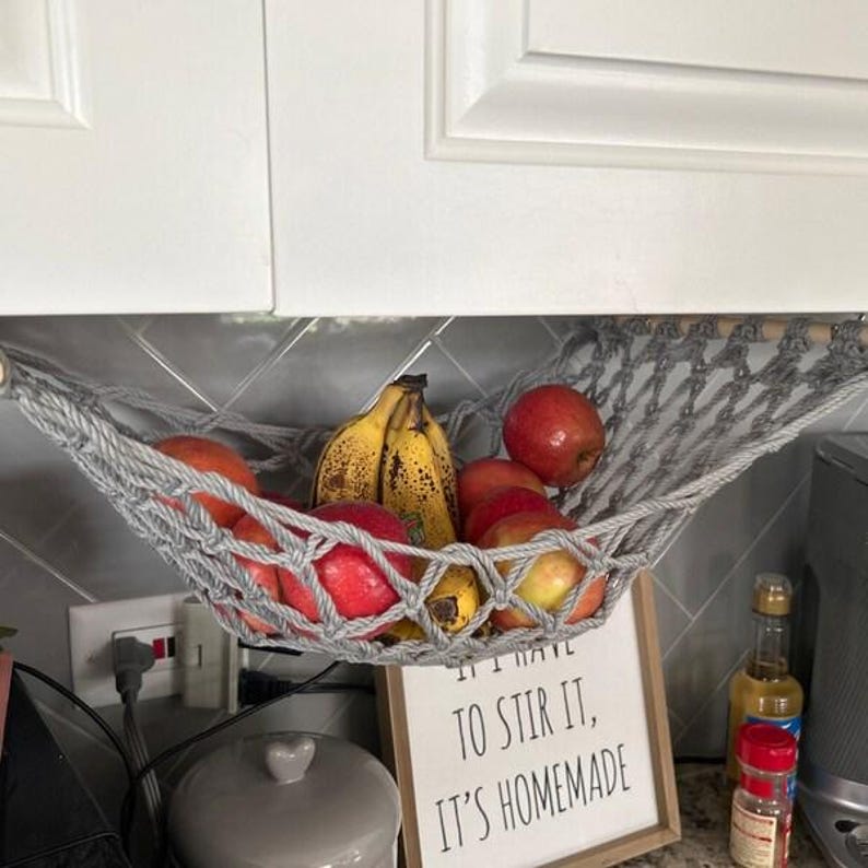 Puede incluir: Una hamaca de frutas de macram&eacute; gris llena de manzanas rojas y pl&aacute;tanos amarillos. La hamaca est&aacute; suspendida debajo de un armario blanco. Un letrero debajo dice: "I HAVE TO STIR IT, IT'S HOMEMADE".