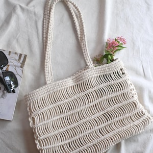 May include: An off-white macrame tote bag featuring a repeating horizontal knotted pattern and two long shoulder straps. The bag contains a small sprig of pink flowers. A pair of black sunglasses and a printed card are visible to the left.